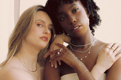 Two models wearing layered Goldmacher moissanite jewelry – featuring tennis chains, pendant necklaces, and stacked rings in gold and silver finishes