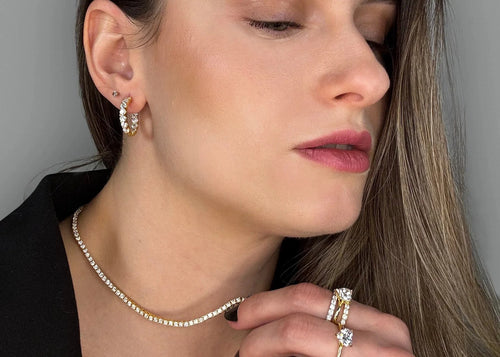 Hands-focused photo showing Goldmacher moissanite heart, pavé, and V-shaped rings, with hoop earrings and tennis chain mobile